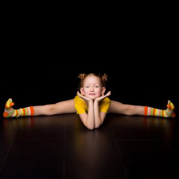 Cute happy young girl gymnast doing cross splits. Happy adorable kid wearing yellow leotard performing rhythmic gymnastic exercise and smiling at camera against black background