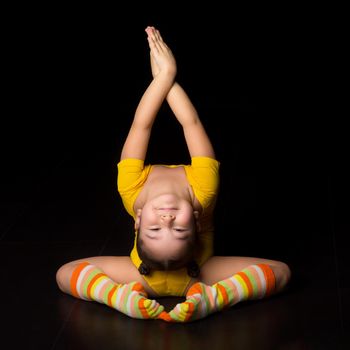 Cute flexible little girl acrobat doing acrobatic exercise. Girl gymnast wearing yellow leotard and striped socks performing back bend pose against isolated black background in studio