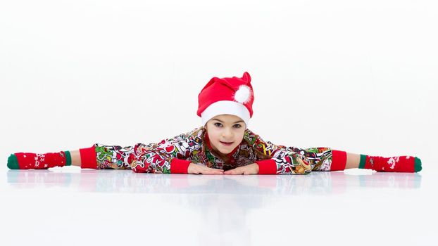 Happy flexible little girl gymnast doing cross splits. Cheerful girl wearing bright colorful overalls, socks and Santa Claus cap performing splits against isolated white background