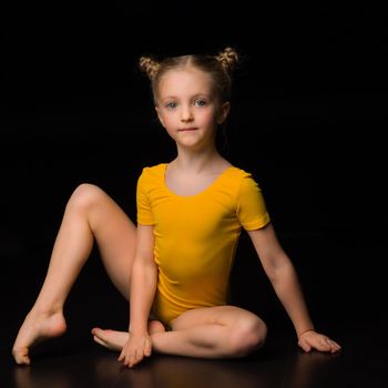 Beautiful blue eyed girl gymnast in yellow leotard. Lovely blonde barefoot kid dancer or acrobat sitting on the floor against isolated black background in studio