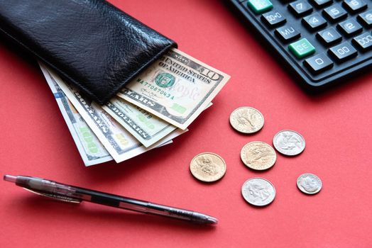 black leather wallet with dollar bills of different denominations, coins, a pen and a calculator on a red background.