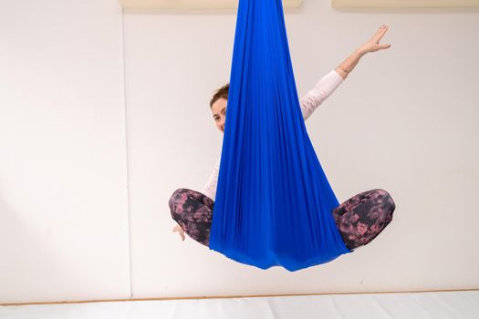 A young woman poses while doing anti-gravity aerial yoga in a blue hammock on a white background