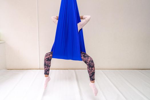 A young woman poses while doing anti-gravity aerial yoga in a blue hammock on a white background