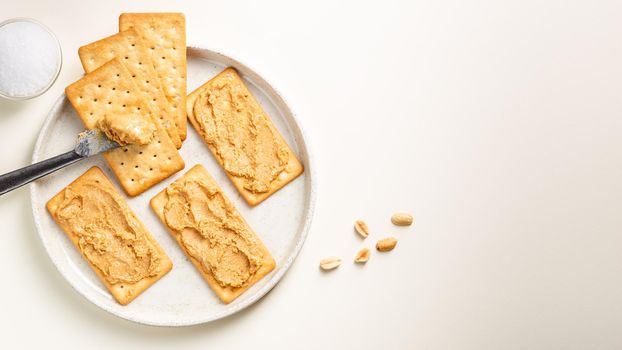Peanut butter spread on soda crackers on the plate seen from above with knife and salt. Above view