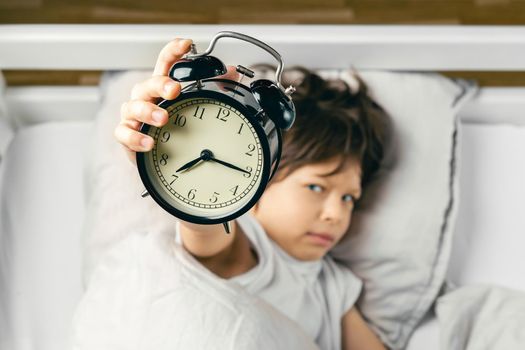 Sleepy grumpy boy is holding an alarm clock that has awaken him up in the morning. Focus on the clock