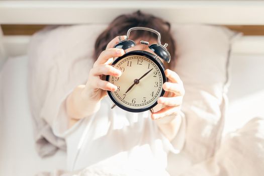 Unrecognizable kid lying in bed and holding a retro style alarm clock. Time to get up in the morning. Focus on the clock