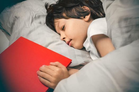 Cute boy peacefully sleeping at night in his bed with a red story book. Kids literature concept. Reading before bed for children