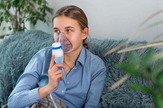Teenage girl makes inhalation with a nebulizer equipment. Sick child holding inhalator in hand and breathes through an inhaler at home. Physical therapy for cold, flu and bronchial asthma