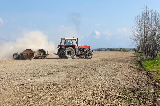 Liptovsky Hradok, Slovakia - April 22nd, 2018: Tractor pulling heavy metal roller, preparing dry field in the spring, dust cloud behind, with mount Krivan, Slovak symbol, in the background.