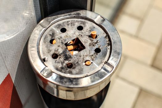 Sun shining on metal ashtray stand outside restaurant / office, with cigarette butts and ash. Smoking ban inside concept.