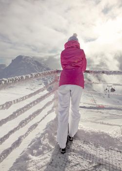 Woman in pink ski jacket leaning on snow covered rail, looking at ski piste, with sun and clouds backlight in background.