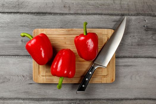 Table top view - three bright red bell peppers, water drops, with chefs knife on chopping board, with rustic gray wood desk under.