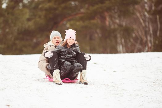 Happy mother with baby girl sitting on sledge and sledding down on snow from hill. Enjoying white winter day at park. Spending time together in weekend