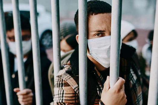 Shot of a young man wearing a mask while stuck behind a gate in a foreign city