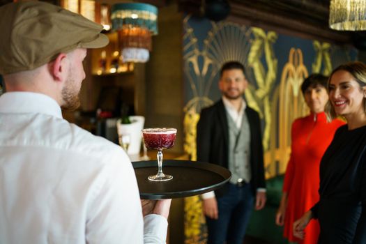 Back view of male waiter in white shirt serving red alcohol cocktail on tray for group of elegant people in luxury bar