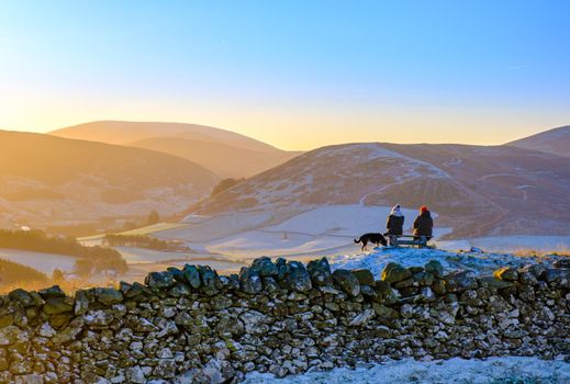 Two Friends Sitting On A Bench In The Beautiful Scottish Countryside After A Hike On A Frosty Morning, With Their Dog