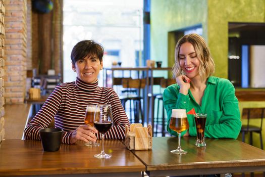 Happy adult women in casual clothes smiling and enjoying alcohol drinks while sitting at table during friends meeting on weekend day in pub