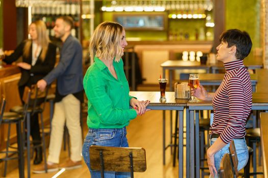 Side view of happy blond woman in casual clothes smiling and listening to girlfriend drinking beer while spending time in pub together