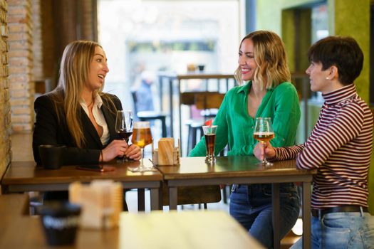 Happy adult women with alcohol drinks smiling and talking while gathering around table during friends meeting in pub