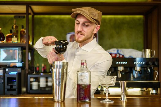 Bearded man in white shirt and cap squeezing lemon juice over shaker with cocktail while working behind counter in pub