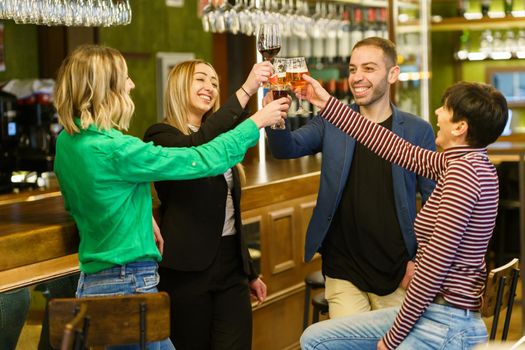 Merry multiethnic adult man and women in casual clothes smiling and clinking glasses of alcohol drinks while resting near counter in weekend in bar