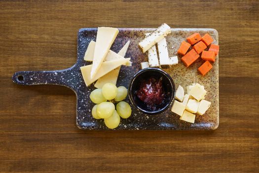 Top view of ripe grapes and pieces of assorted cheese placed on board around bowl with sauce on restaurant table