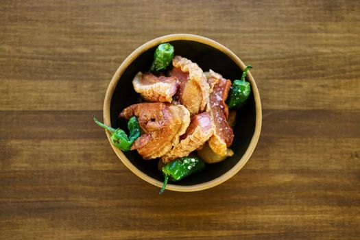 Top view of torreznos, pieces of deep fried, meat with green hot peppers served in bowl on lumber table in restaurant