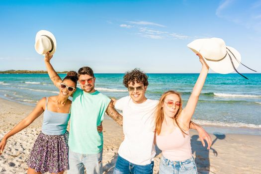 Group of gen z friends embracing to each other looking at camera on the seashore in tropical ocean resort. Happy students enjoying sea vacations together. Carefree people posing for photography