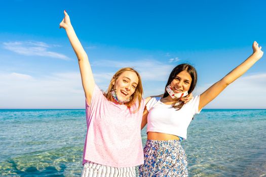 Two happy gen z girls embracing to each other with raised open arms looking at camera wearing funny protective open face mask enjoying sea vacations with crystal sea water and blue sky in background