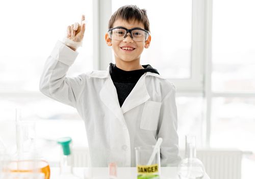 School boy wearing protection glasses during chemistry experiment in elementary science class looking at camera with hand up and smiling. Clever pupil in lab during test