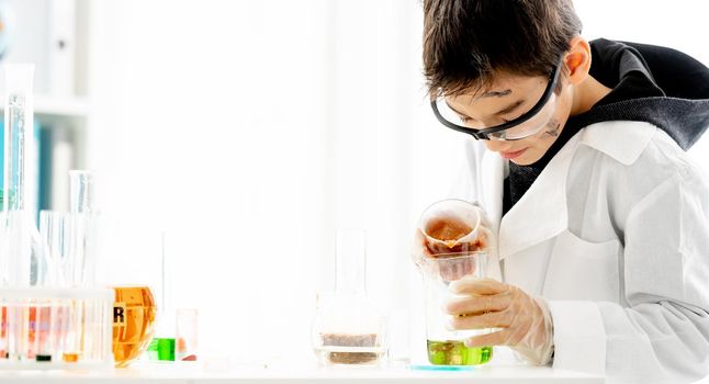 School boy wearing protection glasses mixing liquids in tubes during chemistry experiment in elementary science class. Clever pupil in lab during test