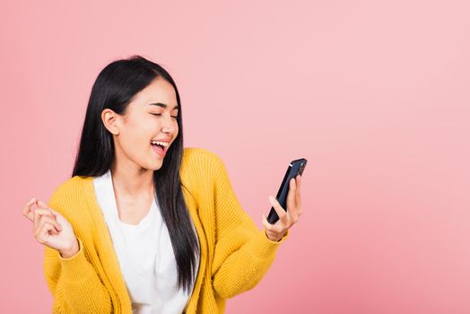 Happy Asian portrait beautiful cute young woman excited laughing holding mobile phone, studio shot isolated on pink background, female using funny smartphone making winner gesture