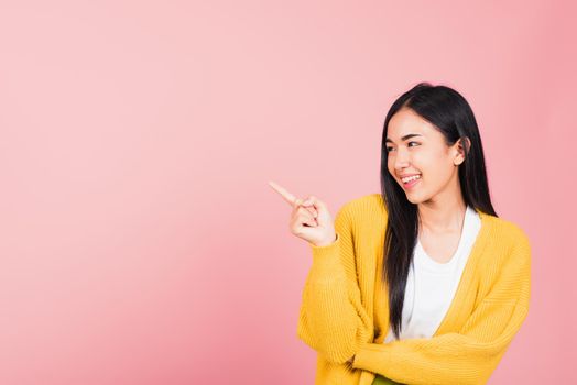 Asian happy portrait beautiful cute young woman standing pointing finger side away presenting product looking to away side, studio shot isolated on pink background with copy space