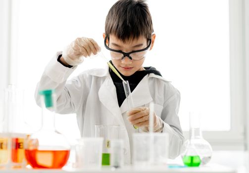 School boy wearing protection glasses doing chemistry experiment in elementary science class. Clever pupil with tubes measuring liquids in laboratory classroom