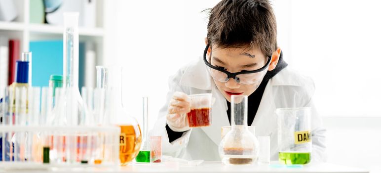 School boy with dirty face wearing protection glasses looking at liquid in glass during chemistry experiment in elementary science class and smiling. Clever pupil in lab during test