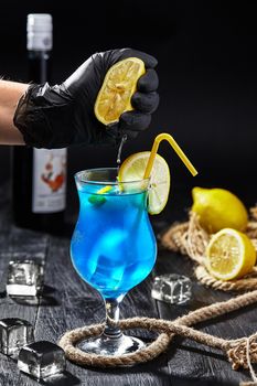 Male hand in black rubber glove squeezing fresh lemon into glass with Blue Lagoon. Preparation of refreshing and sweetish cocktail based on vodka with addition of Blue Curacao liqueur and lemonade