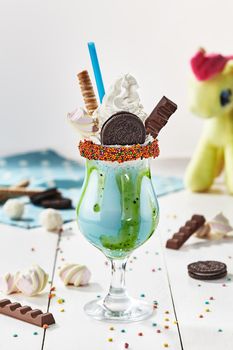 Delicate milkshake topped with whipped cream, waffle roll, chocolate cookie, marshmallow and candy bar in glass garnished with colorful sprinkles. Kids sweet party menu concept