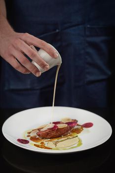 Closeup of male hand pouring white sauce on plate with juicy baked duck breast with slices of beetroot and daikon. Chef preparing dish for serving