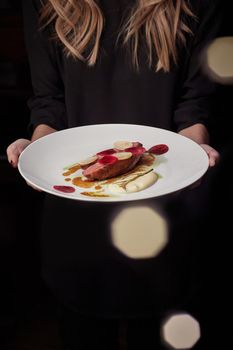 Young female hands holding plate with delicious juicy baked duck breast with thin slices of beetroot and daikon and bechamel sauce, dark background with dotted bokeh effect