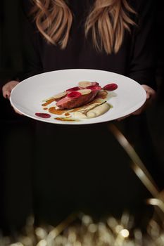 Young woman in black clothes holding plate with roasted duck breast garnished with thin slices of daikon and beetroot seasoned with bechamel sauce. Waitress serving guests