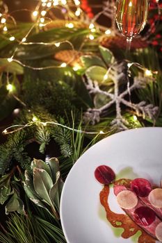 Glass of champagne and plate with delicious baked duck breast served with thin slices of daikon and beetroot on festive New Year dining table decorated with fir branches and garland lights