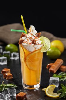 Delicious refreshing lemonade with lime and orange slices topped with delicate whipped cream, sweet toffees, caramel sauce and colorful sprinkles on black background. Unusual mix of drink and dessert