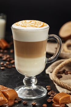 Glass cup of delicious latte macchiato with milk foam topped with caramel syrup on black background with coffee beans and sweet toffees