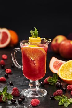 Aromatic fruit and berry tea from ripe raspberry and currant served with slices of orange, grapefruit and fresh mint leaves in glass on black background. Healthy vitamin drinks concept