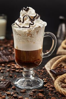 Delicious mochaccino with espresso layer and fluffy cloud of whipped cream topped with chocolate icing in glass cup on black table with coffee beans and pieces of chocolate