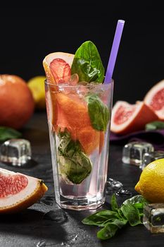 Refreshing citrus drink with pink grapefruit and basil leaves served in glass with crushed ice and straw on black background. Summer nonalcoholic party concept