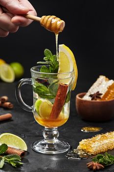 Male hand adding honey with dipper in freshly brewed oriental style tea with lime and lemon slices, cinnamon stick, anise star and aromatic mint leaves in glass on black background