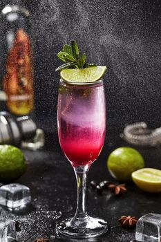 Refreshing alcoholic cocktail based on sambuca and white rum with lime juice, soda and currant syrup served in glass garnished with lime slice and mint leaves on black background in water spray