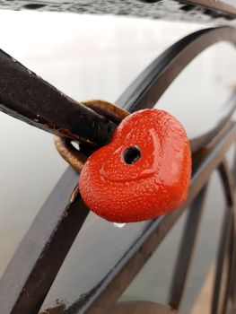 Red lock in the shape of a heart on the metal fence of the city embankment. The lock is metal hinged.