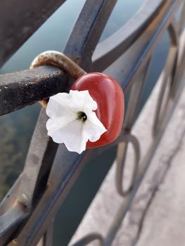 Red castle in the shape of a heart on the metal fence of the city embankment with a white flower. Metal padlock with white flower.